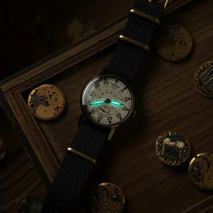 May include: A vintage-style wristwatch with a black nylon strap and gold-tone hardware. The watch face is off-white with black numerals and hands, and a luminous green glow. The watch is set on a wooden surface with several gold-colored metal objects.