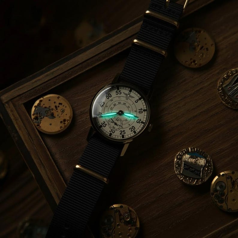 May include: A vintage-style wristwatch with a black nylon strap and gold-tone hardware. The watch face is off-white with black numerals and hands, and a luminous green glow. The watch is set on a wooden surface with several gold-colored metal objects.