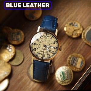 May include: A gold wristwatch with a cream face and a blue leather strap. The watch face has numbers and sub-dials. The text "BLUE LEATHER" is in a blue rectangle at the top. The watch is surrounded by antique coins.
