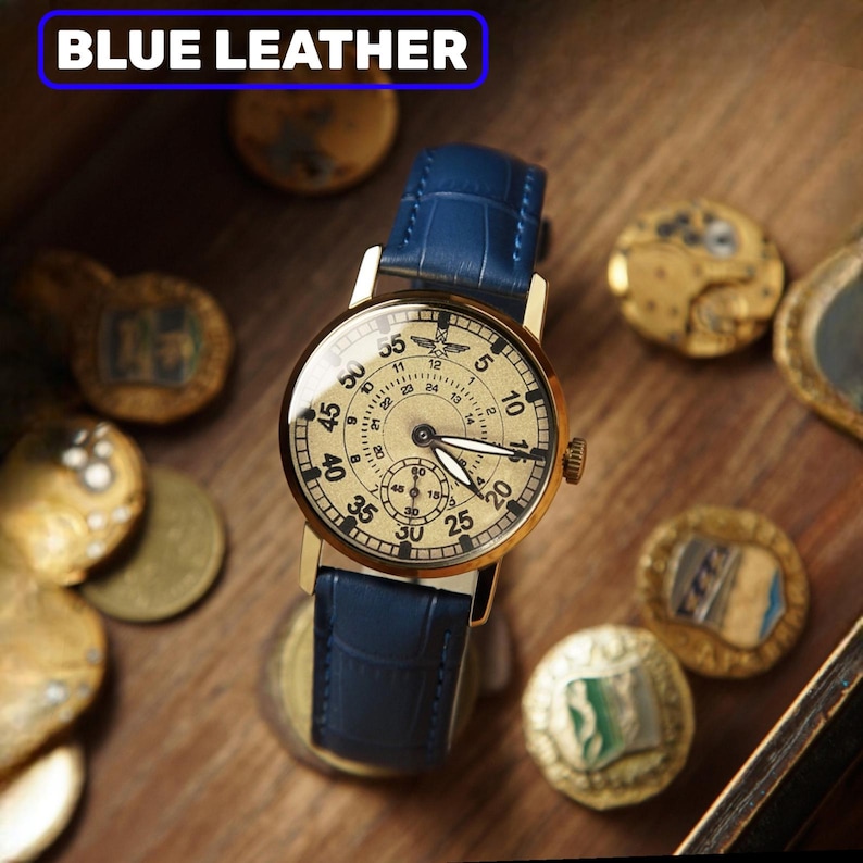 May include: A gold wristwatch with a cream face and a blue leather strap. The watch face has numbers and sub-dials. The text "BLUE LEATHER" is in a blue rectangle at the top. The watch is surrounded by antique coins.