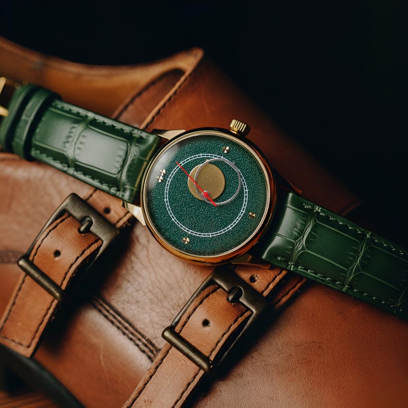 May include: A gold-toned wristwatch with a green face and a green leather band rests on a brown leather shoe. The watch face features a circular design with a red second hand. The watch band has a crocodile texture.