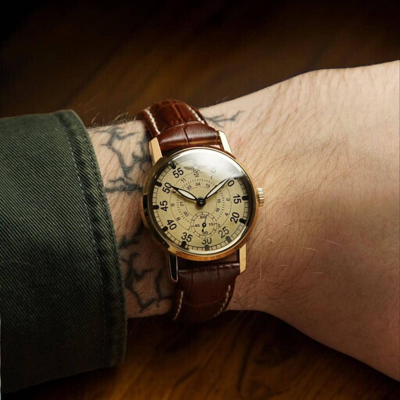 Puede incluir: Un reloj de pulsera dorado con esfera color crema y correa de cuero marr&oacute;n. El reloj tiene m&uacute;ltiples subesferas y un cristal transparente. La correa se sujeta con una hebilla. El reloj est&aacute; en una mu&ntilde;eca.