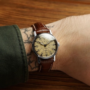 Puede incluir: Un reloj de estilo vintage con caja plateada y esfera de color beige claro. El reloj presenta n&uacute;meros y agujas negros, y una correa de cuero marr&oacute;n. El reloj se lleva en la mu&ntilde;eca.