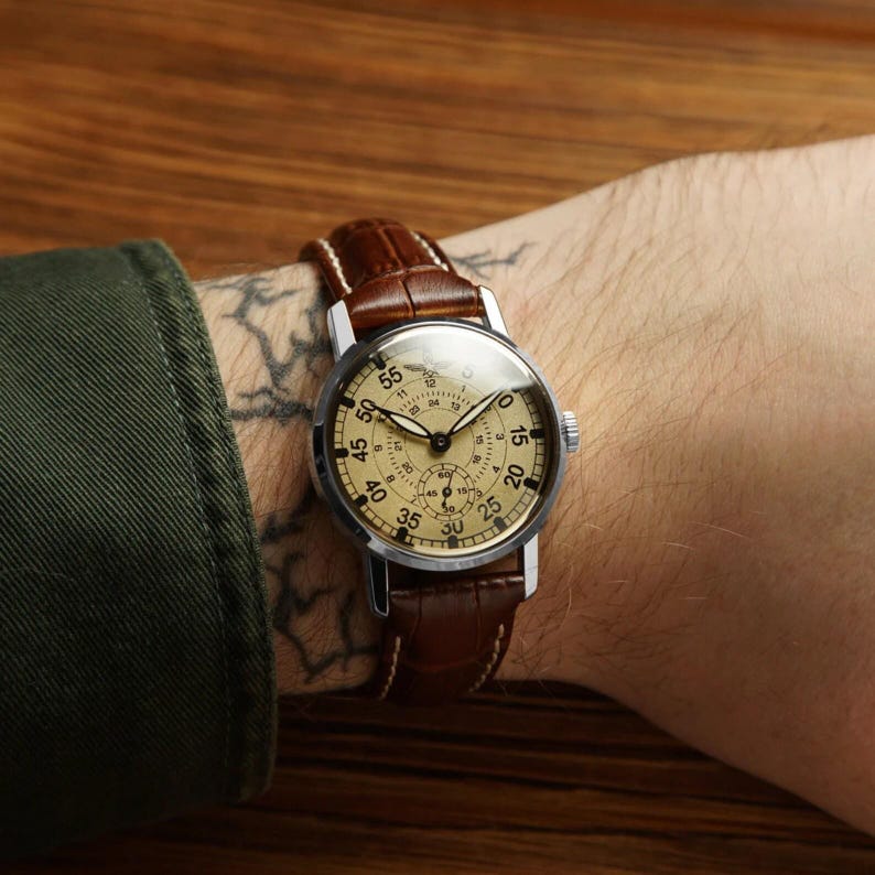 Puede incluir: Un reloj de estilo vintage con caja plateada y esfera de color beige claro. El reloj presenta n&uacute;meros y agujas negros, y una correa de cuero marr&oacute;n. El reloj se lleva en la mu&ntilde;eca.