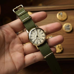 May include: A vintage-style wristwatch with a cream-colored face and gold-tone case. The watch features black numerals and hands, and a small subdial. It has a green nylon strap with gold-tone hardware. The watch is approximately 1.5 inches in diameter.