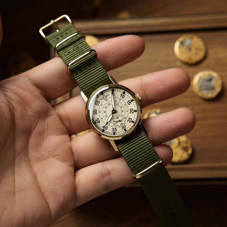 May include: A vintage-style wristwatch with a cream-colored face and gold-tone case. The watch features black numerals and hands, and a small subdial. It has a green nylon strap with gold-tone hardware. The watch is approximately 1.5 inches in diameter.