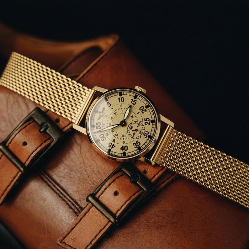 Peut inclure: Une montre-bracelet dor&eacute;e avec un bracelet en maille repose sur une surface en cuir marron. Le cadran de la montre est cr&egrave;me avec des chiffres et des aiguilles noirs. La montre est associ&eacute;e &agrave; une chaussure en cuir marron avec deux boucles.