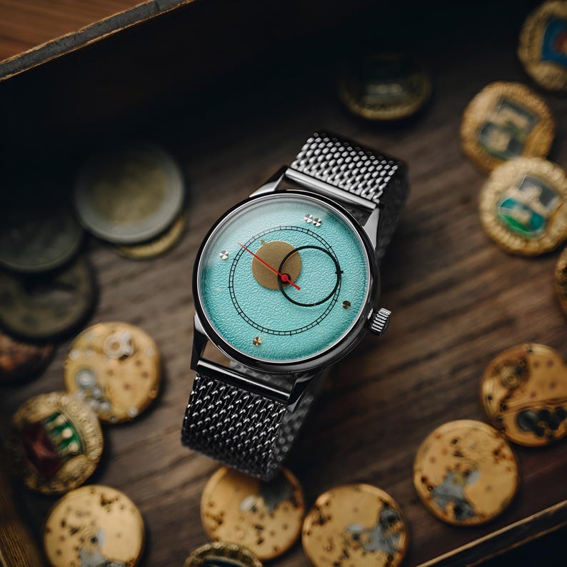 Op de afbeelding: Een zilveren polshorloge met een mesh band en een lichtblauwe wijzerplaat. De wijzerplaat heeft een goudkleurige cirkel, een zwarte cirkel en een rode wijzer. Het horloge ligt op een houten oppervlak met munten.
