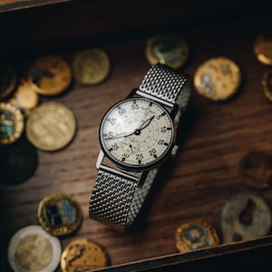 Puede incluir: Un reloj de estilo vintage con una correa de malla plateada y una esfera redonda de color crema. El reloj presenta agujas y n&uacute;meros negros, con una esfera secundaria m&aacute;s peque&ntilde;a. El reloj est&aacute; sobre una superficie de madera, rodeado de monedas de oro.