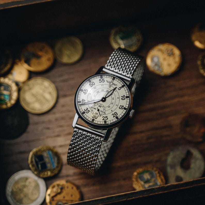 Puede incluir: Un reloj de estilo vintage con una correa de malla plateada y una esfera redonda de color crema. El reloj presenta agujas y n&uacute;meros negros, con una esfera secundaria m&aacute;s peque&ntilde;a. El reloj est&aacute; sobre una superficie de madera, rodeado de monedas de oro.