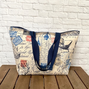 May include: A large canvas tote bag with navy blue handles. The bag is printed with a vintage postage stamp design in shades of blue, red, and black. The text on the bag reads "LE", "TE POST", "sole est extur", "re", "12.1008.5-6%", "25", "Ja ipie", "Quend Amp", "OSTA", "1261", "PAR", and "PARTEL".