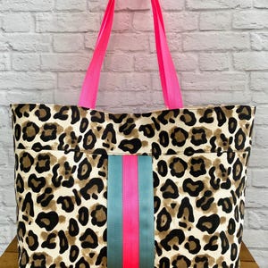May include: A tote bag featuring a beige, brown, and black leopard print pattern. The bag has two bright neon pink shoulder straps and a vertical center stripe detail consisting of two teal bands flanking a central neon pink stripe.