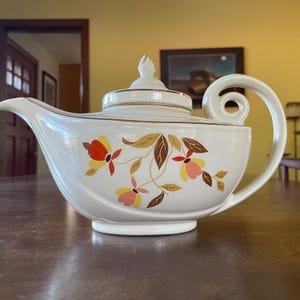 May include: A white teapot with a gold trim and a floral design in shades of orange, yellow, pink, and brown. The teapot has a curved spout and a handle that curls around the top.