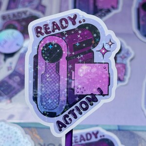Peut inclure: Un autocollant holographique représentant une caméra vidéo vintage violette et bleue avec le texte READY.. ACTION ! Le design comprend des accents d'étoiles scintillantes et une bordure découpée, affichant une palette de couleurs violet et indigo.