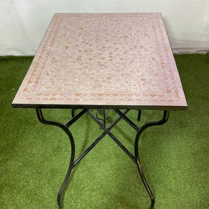 Peut inclure: Table d'extérieur rectangulaire avec un dessus en mosaïque dans les tons de rose et de beige. La table a une base en fer forgé noir avec des pieds incurvés. La table est posée sur une surface verte.