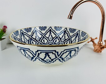 Può includere: Un lavabo decorativo in ceramica con un motivo geometrico blu e bianco e un bordo dorato. Un rubinetto in rame è posizionato a destra. Una piccola pianta in vaso con fiori rosa è a sinistra.
