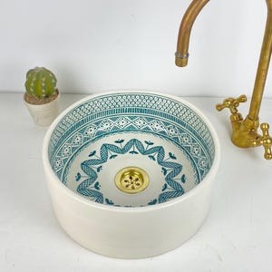 Puede incluir: Un lavabo redondo de cerámica blanca con un interior estampado en blanco y turquesa. Un grifo de latón está colocado encima del lavabo. Un pequeño cactus en maceta está a la izquierda.
