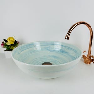 Könnte beinhalten: Ein weißes Keramik-Waschbecken mit blauem Wirbelmuster. Das Waschbecken ist mit einem kupferfarbenen Wasserhahn kombiniert. Links vom Waschbecken befindet sich eine kleine Topfpflanze mit gelben Blüten.