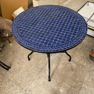 Pode incluir: Mesa redonda de exterior com tampo de mosaico azul. A mesa tem uma estrutura de metal preto e três pernas curvas. O padrão de mosaico apresenta pequenas telhas geométricas. Adequada para pátio ou jardim. Diâmetro de aproximadamente 76 cm.