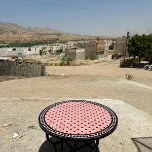 Peut inclure: Une table ronde en métal noir avec un plateau en mosaïque rose et noir. La table est posée sur une surface en terre avec une vue sur une petite ville en arrière-plan.