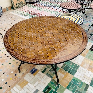 Puede incluir: Una mesa redonda de mosaico con base de metal oscuro. La tapa presenta un patrón de pequeñas baldosas de forma irregular en tonos marrones y beige. La mesa tiene aproximadamente 100 cm de diámetro.
