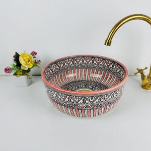Puede incluir: Un lavabo decorativo de cerámica con un diseño de patrones rojos y blancos. La cuenca presenta un patrón geométrico detallado y un borde rosa. Un grifo dorado es visible en el fondo, lo que sugiere un entorno de baño o aseo.