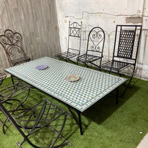 Peut inclure: Un ensemble de patio en fer forgé noir avec une table rectangulaire et six chaises assorties. La table a un dessus en mosaïque dans les tons de vert et de blanc. Les chaises ont des dossiers décoratifs.