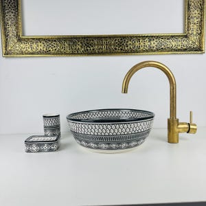 Puede incluir: Un lavabo de baño de cerámica blanca con un patrón geométrico negro. El lavabo es redondo y tiene un grifo de latón. Hay dos pequeños recipientes de cerámica blanca con patrones geométricos negros al lado del lavabo.