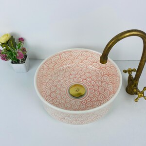 May include: A round ceramic sink with a pink and white patterned design. The sink has a gold-colored drain and is paired with a vintage-style brass faucet. A small potted plant with yellow and pink flowers is in the background.