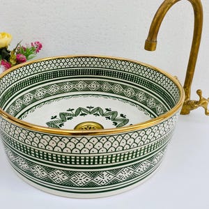 May include: A round ceramic sink with a decorative green and white pattern and a gold rim. A gold tap is positioned to the right of the sink. The sink has a decorative floral design in the centre.