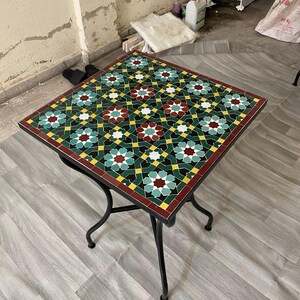 Può includere: Tavolo quadrato in mosaico con base in ferro battuto nero. Il piano presenta un motivo geometrico in verde, rosso, giallo e bianco. Il tavolo misura circa 61 cm quadrati.