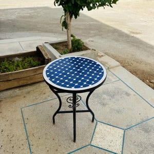 May include: A round outdoor side table with a mosaic tile top. The tabletop features a blue and white geometric pattern. The table has a black metal frame with decorative scrollwork and three legs. The table is approximately 24 inches in diameter.