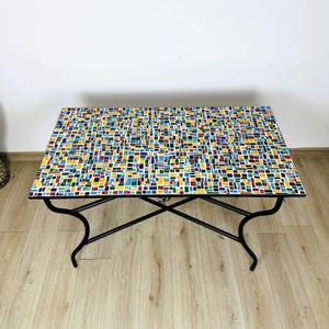 Peut inclure: Une table rectangulaire avec un cadre en métal noir et un plateau en mosaïque coloré. Les carreaux sont dans des tons de jaune, bleu, vert, rouge et blanc.