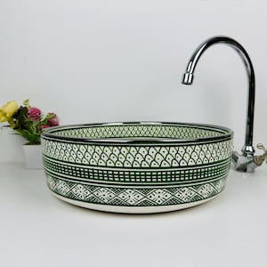 Könnte beinhalten: Ein runder, weißer Keramikwaschtisch mit einem grün-weißen geometrischen Muster. Über dem Waschbecken befindet sich ein verchromter Wasserhahn. Links vom Waschbecken steht eine kleine Vase mit Blumen.