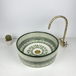 Puede incluir: Lavabo redondo de cerámica con un patrón geométrico verde y blanco. El lavabo tiene un diseño floral central y un grifo de latón. A la izquierda del lavabo hay un pequeño cactus en una maceta blanca.