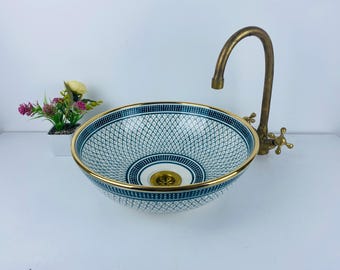 May include: A white ceramic vessel sink with a blue and gold geometric pattern. The sink has a gold rim and a drain in the centre. A brass faucet is mounted to the wall behind the sink.