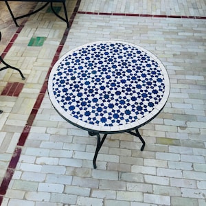 Peut inclure: Une table ronde en métal noir avec un plateau en carrelage bleu et blanc. Le plateau de la table présente un motif géométrique d'étoiles bleues sur fond blanc.