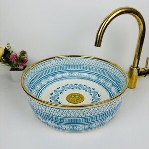 May include: A white ceramic sink with a blue and gold geometric pattern. The sink has a gold rim and a gold drain.