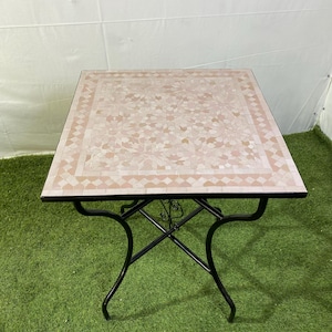 May include: Square outdoor table with a mosaic tile top in shades of pink and beige. The table has a black metal frame with curved legs. The table is approximately 24 inches square.