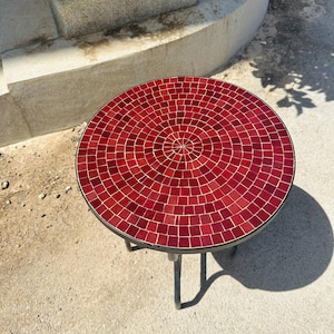 May include: A round red mosaic table with a black metal frame. The table top is made of small red tiles arranged in a circular pattern.