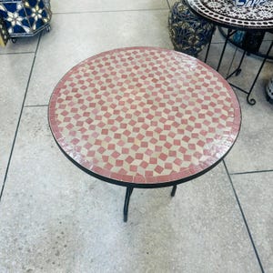 Könnte beinhalten: Kleiner, runder Beistelltisch mit Mosaikfliesen. Die Tischplatte hat ein Muster aus abwechselnd rosa und cremefarbenen Quadraten. Der Tisch hat ein schwarzes Metallgestell und drei gebogene Beine. Der Tisch hat einen Durchmesser von ca. 46 cm.