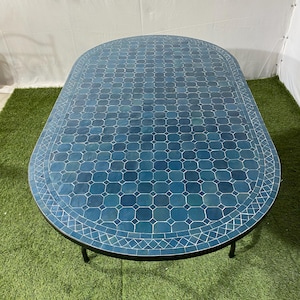 Può includere: Tavolo ovale da esterno con piano a mosaico blu. Il motivo a mosaico è geometrico. Il tavolo ha una struttura e gambe in metallo nero. È posizionato su una superficie verde. Dimensioni approssimative: 150 cm x 90 cm.