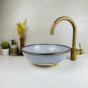 Può includere: Un lavabo in ceramica bianca con un motivo geometrico blu e bianco. Il lavabo ha un bordo dorato e una base dorata. Un rubinetto dorato è montato sopra il lavabo.