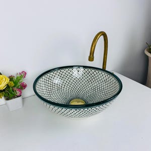 Può includere: Un lavandino in ceramica bianca con un motivo geometrico verde e bianco. Il lavandino è rotondo e ha un piccolo foro di scarico al centro. Un rubinetto dorato è montato sul muro dietro il lavandino.