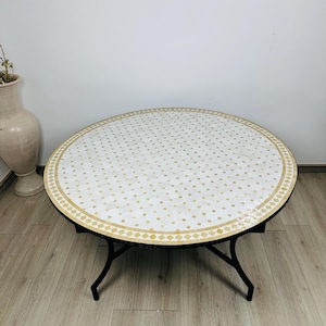 May include: A round, white mosaic coffee table with a gold border and black iron legs. The table is on a light wood floor.