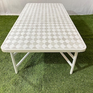 May include: A rectangular folding table with a white frame and legs. The tabletop features a repeating pattern of gray and white curved shapes. The table is set on a green surface.
