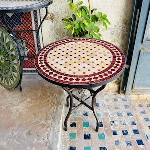 Può includere: Un tavolo rotondo in mosaico con una struttura in metallo scuro. Il piano del tavolo presenta un motivo di piccole piastrelle beige e bordeaux. Il tavolo è sostenuto da quattro gambe curve. Un altro tavolo con un piano in mosaico verde è parzialmente visibile.