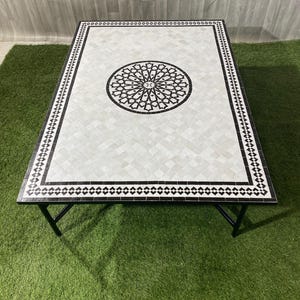 May include: A rectangular coffee table with a mosaic tile top. The table has a white and black geometric pattern with a central circular design. It has a black metal frame and legs.