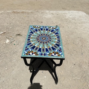 May include: A small, rectangular side table with a mosaic tile top. The tile design features a symmetrical pattern in shades of blue, teal, brown, and gold. The table has a black metal frame and legs.