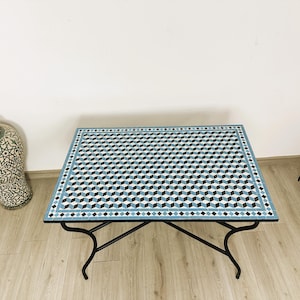 Peut inclure: Une table carrée en mosaïque avec un motif géométrique bleu et blanc. La table a un cadre en métal noir et est posée sur un sol en bois.